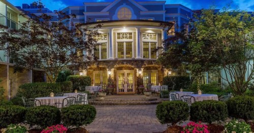 Elegant building with large windows, surrounded by greenery and outdoor seating, illuminated at dusk.