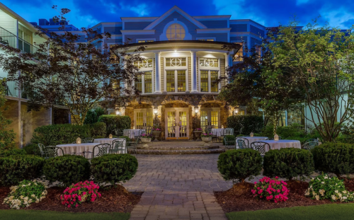 A charming building with elegant architecture, surrounded by lush greenery and colorful flowers, illuminated at dusk.