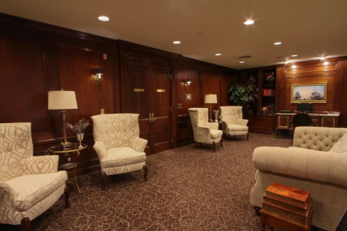 A cozy, elegantly furnished room with plush chairs, a desk, and warm wood paneling. Soft lighting enhances the ambiance.