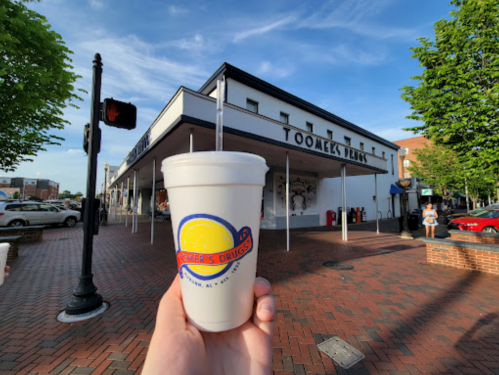 A hand holds a drink cup in front of Toomey's Drugs, a brick-paved street with trees and a blue sky in the background.
