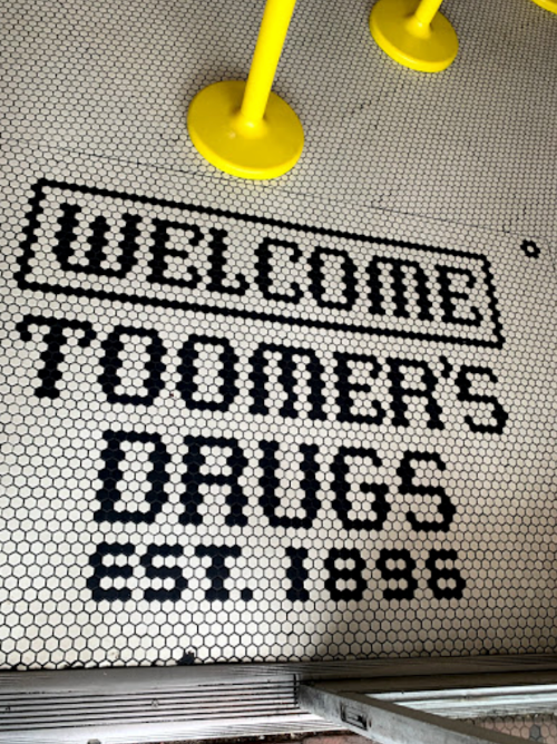 Black and white tiled floor with "Welcome Toomer's Drugs EST. 1896" in bold lettering. Yellow poles in the background.