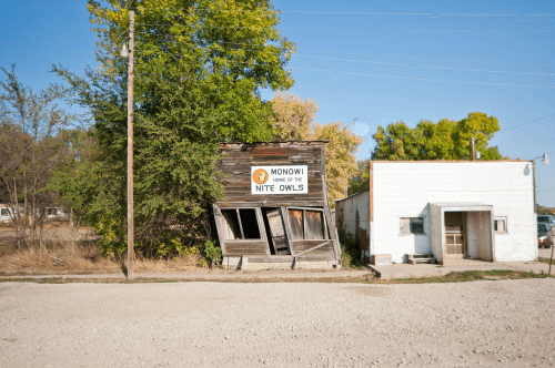 A leaning wooden building with a sign reading "Monowi, Home of the Nite Owls," next to a white structure.