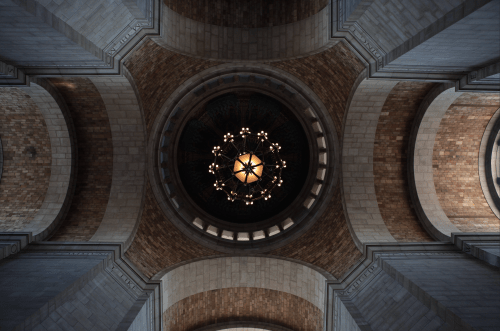 A view of a grand ceiling with a decorative chandelier and arched stone architecture.