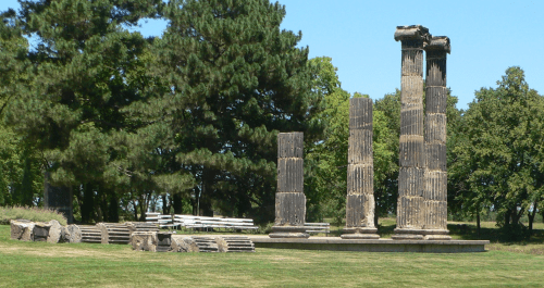 Ancient stone columns and ruins surrounded by trees in a grassy area under a clear blue sky.