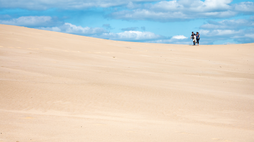 Two people walking on a vast sandy dune under a blue sky with fluffy clouds.