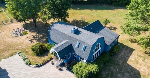 Aerial view of a modern gray house surrounded by greenery and a circular fire pit area in the yard.