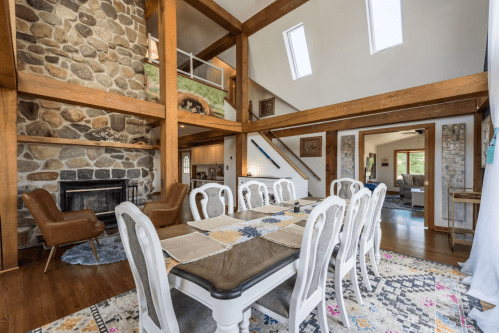 A spacious dining area with a large table, stone fireplace, and wooden beams, leading to a cozy living space.