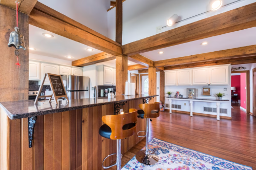 A modern kitchen with wooden beams, a granite countertop, bar stools, and white cabinetry, featuring warm lighting.
