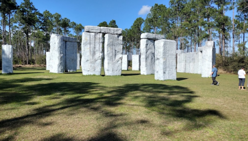 A replica of Stonehenge made of large, gray stones in a grassy area surrounded by trees. Two people are walking nearby.