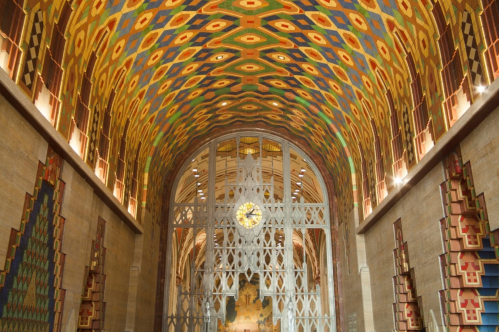 Vibrant, patterned ceiling of a grand hall with decorative walls and a large clock behind a metal gate.