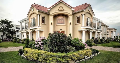 A grand, two-story house with a landscaped garden and blooming flowers, set against a cloudy sky.