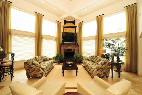 Bright living room with large windows, two patterned sofas, a coffee table, and a fireplace, surrounded by plants.