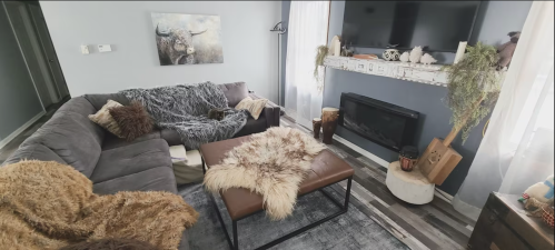 Cozy living room with a gray sofa, fur throws, a coffee table, and a fireplace, decorated with plants and artwork.
