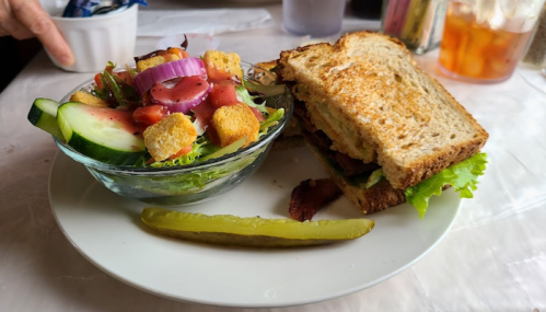 A plate with a sandwich on whole grain bread, a side salad with croutons, and a pickle.