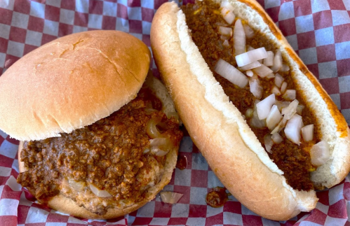 Two sandwiches on a red and white checkered paper: one with chili and onions on a bun, the other in a hot dog roll.