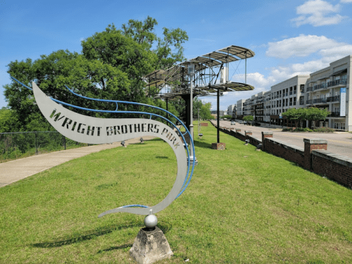 Sign for Wright Brothers Park with a model of an airplane in the background and a clear blue sky.
