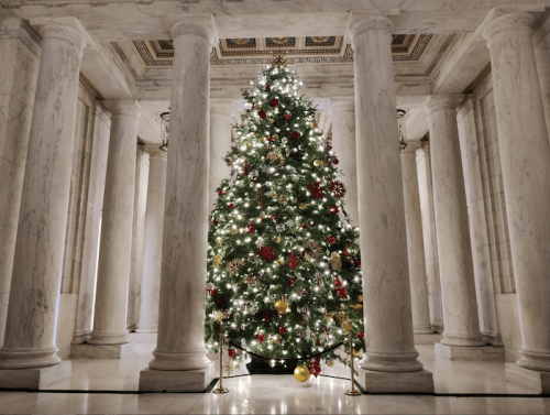 A beautifully decorated Christmas tree with lights and ornaments stands in a grand marble hall surrounded by columns.