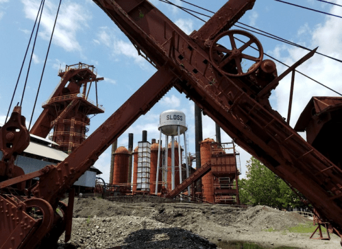 Rusty industrial structures and a water tower labeled "SLOSS" against a blue sky with scattered clouds.