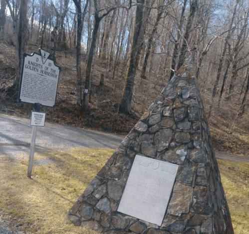 A stone monument with a plaque beside a historical marker in a wooded area, indicating the Knights of the Golden Horseshoe.