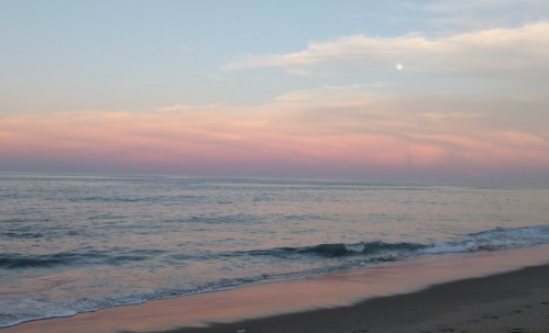 A serene beach scene at sunset, with soft pink clouds and a calm ocean reflecting the colors.