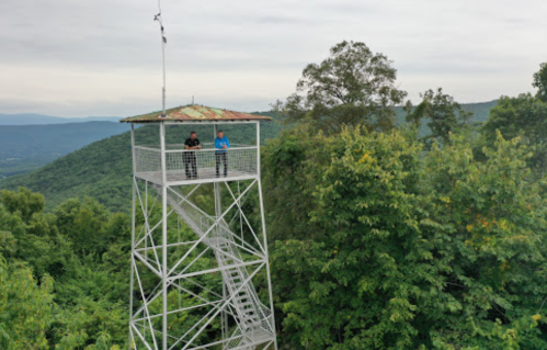 Two people stand on a lookout tower surrounded by lush green trees and mountains under a cloudy sky.
