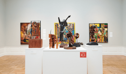 A gallery display featuring various sculptures and artworks on a white pedestal, with colorful paintings in the background.