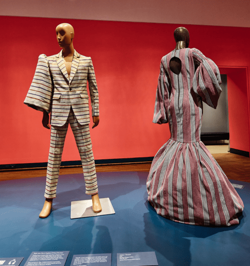 Two mannequins display striking fashion designs: a striped suit and a dramatic gown with puffed sleeves.