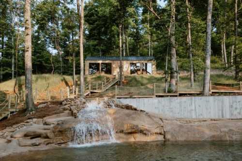 A rustic cabin surrounded by trees, with a small waterfall and a wooden deck by a serene body of water.