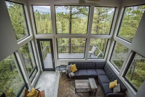 Modern living room with large windows overlooking a forest, featuring a sectional sofa and a balcony.