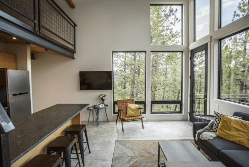 Modern living space with large windows, a cozy sofa, and a kitchen bar overlooking a forested view.