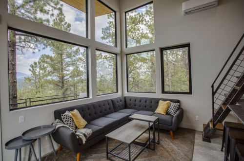 Modern living room with large windows, a gray sectional sofa, and a coffee table, surrounded by trees and mountains.