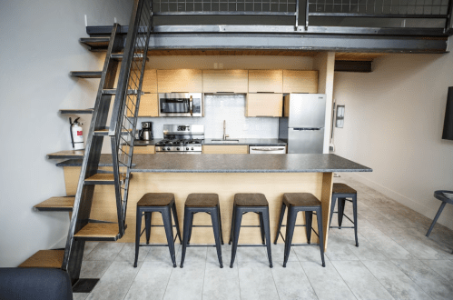 Modern kitchen with wooden cabinets, stainless steel appliances, and a staircase leading to an upper level. Four stools at the bar.