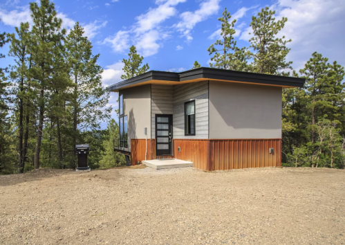 Modern house with a mix of wood and gray siding, surrounded by trees and a gravel area under a blue sky.