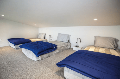 Cozy loft space with four beds, each with gray blankets and blue comforters, under soft lighting.
