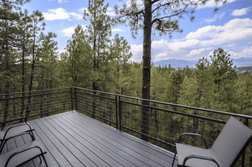 A wooden deck with two chairs overlooks a lush forest of tall pine trees under a partly cloudy sky.