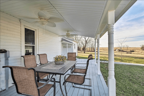 A porch with a table and chairs, featuring a flower centerpiece, overlooking a grassy area and distant fields.