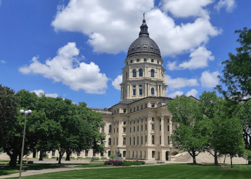 A grand building with a dome, surrounded by trees and blue skies, likely a state capitol or government structure.