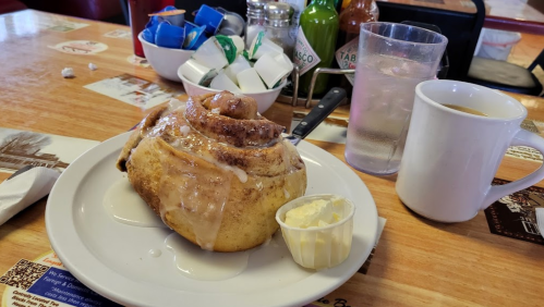 A cinnamon roll with icing on a plate, accompanied by butter, a cup of coffee, and condiments on a wooden table.