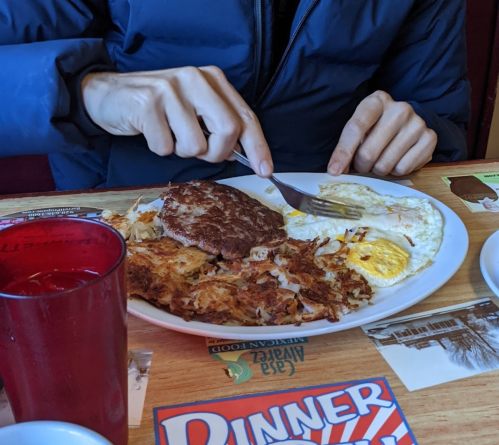 A person cuts into a plate of breakfast, featuring eggs, hash browns, and a sausage patty, with a red drink nearby.