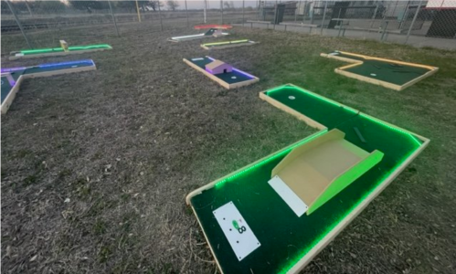 A mini-golf course with illuminated holes and obstacles, set in an outdoor area with grass and a fence in the background.