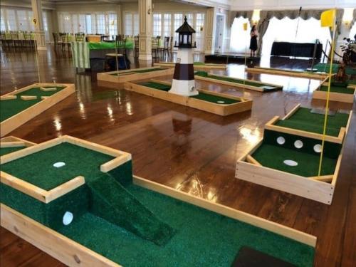 A mini-golf setup indoors with green turf, wooden frames, and a lighthouse feature in a spacious venue.