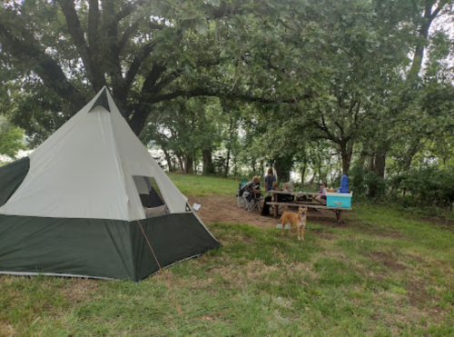 A tent set up in a grassy area with trees, a picnic table, and people gathered nearby, along with a dog.