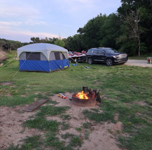 A campsite with a blue and gray tent, a fire pit with flames, and an SUV parked nearby on grassy ground.
