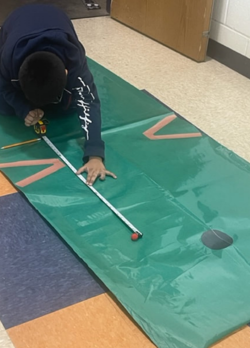 A person measures a green mat with a tape measure, using a pencil to mark measurements on the surface.
