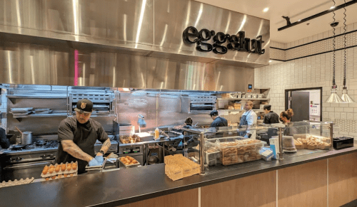 A busy kitchen at Eggslut with chefs preparing food and a display of sandwiches and eggs in the foreground.