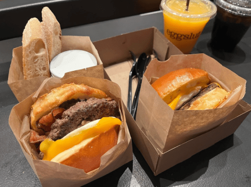 Two burgers in brown paper boxes, one with bacon and cheese, alongside a side of sauce and drinks.