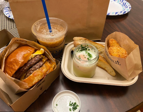 A burger with two patties, a drink, a dessert in a jar, and a side of bread on a table.