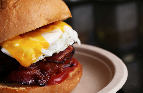 A close-up of a sandwich with a toasted bun, crispy bacon, a fried egg, melted cheese, and ketchup on a paper plate.