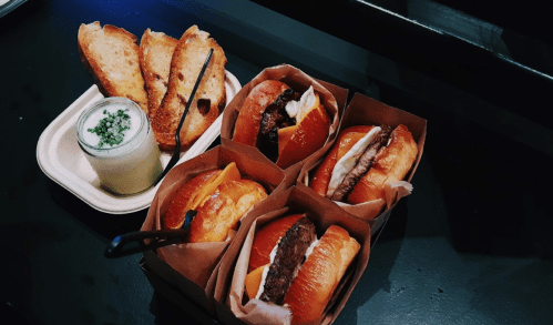 A platter featuring four gourmet burgers in paper containers, alongside toasted bread and a small dish of creamy sauce.
