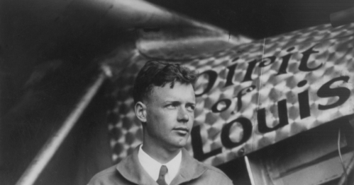 A young man stands in front of an airplane with "Spirit of Louis" written on it, wearing a suit and tie.